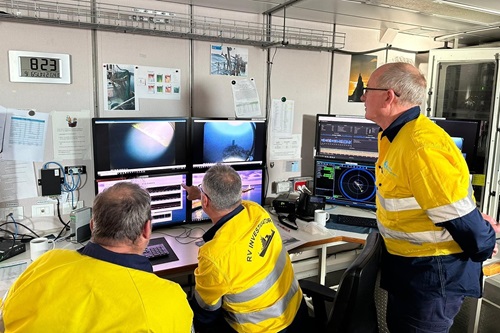 Three people in yellow hi-vis shirts look at a bank of computer screens.